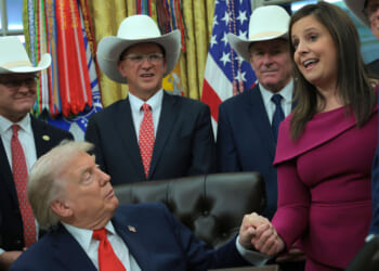Donald Trump holds hands with Rep. Elise Stefanik (R-NY) in the Oval Office with other officials gathered around.