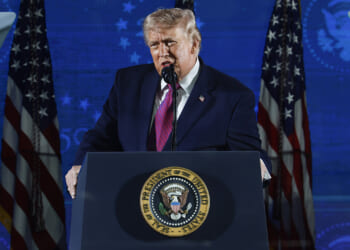 Trump stands at a podium with the presidential seal, speaking into a microphone about the dangers of communism. Multiple American flags are displayed behind him against a blue backdrop.
