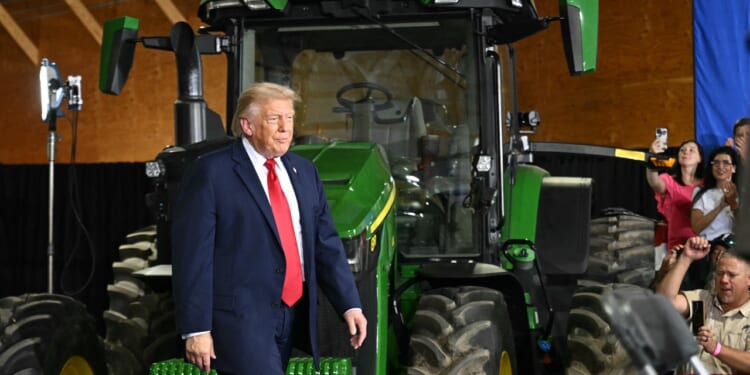 Trump walks past a large green tractor indoors, while people nearby watch and take photos.