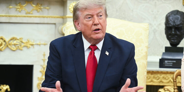A man in a navy suit and red tie sits on a pale yellow chair, gesturing with his hands, in an ornate room with gold details and a dark bust in the background.