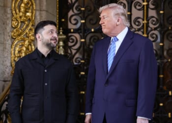 Two men in formal attire stand side by side in front of an ornate gate, looking at each other and smiling. One wears a black jacket, the other a navy suit and blue striped tie.