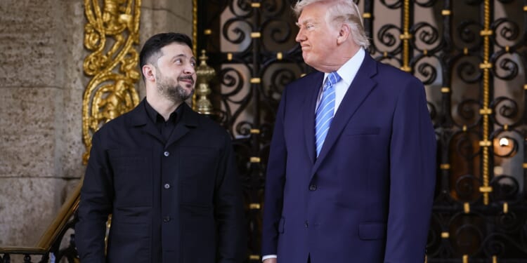Two men in formal attire stand side by side in front of an ornate gate, looking at each other and smiling. One wears a black jacket, the other a navy suit and blue striped tie.