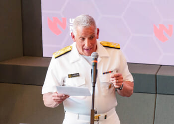 A man in a white military uniform speaks into a microphone while holding papers. Behind him is a screen with the Nebraska Medicine and UNMC logos.