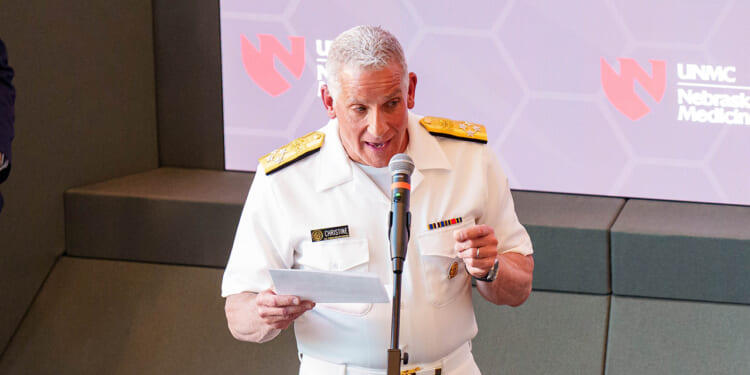 A man in a white military uniform speaks into a microphone while holding papers. Behind him is a screen with the Nebraska Medicine and UNMC logos.