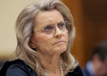 Finland Christian Member of Parliament Paivi Rasanen, an older woman with long gray hair, glasses, and a serious expression sits at a table, wearing a dark outfit and a pearl necklace, with blurred people and a wooden background behind her.