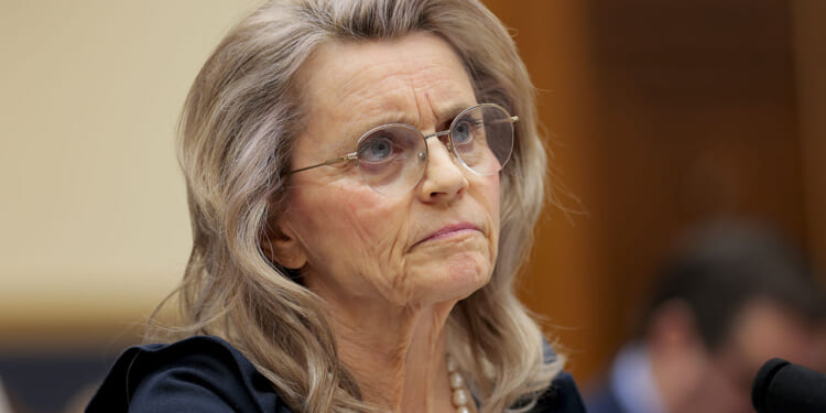 Finland Christian Member of Parliament Paivi Rasanen, an older woman with long gray hair, glasses, and a serious expression sits at a table, wearing a dark outfit and a pearl necklace, with blurred people and a wooden background behind her.