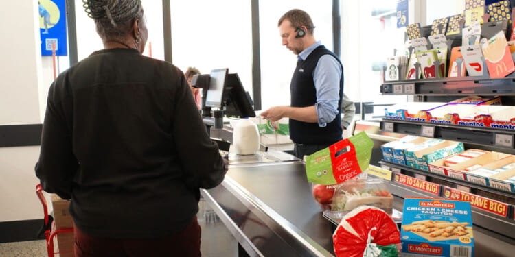 A cashier scans groceries, including bread and cheese, while a woman waits at the register in a brightly lit supermarket.
