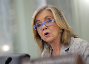A woman with shoulder-length blonde hair, blue glasses, and a plaid blazer speaks, appearing to be at a formal meeting or hearing.