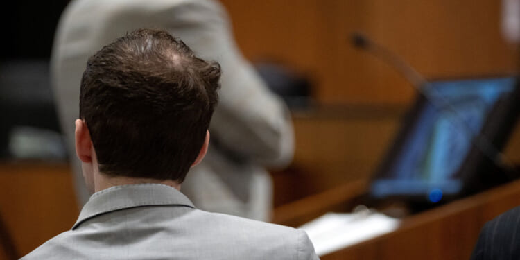A view from behind of Tyler Robinson, in gray jacket, watching prosecutor present case against him.