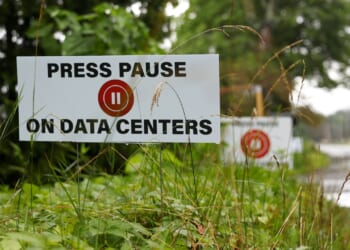 A sign among grass reads PRESS PAUSE ON DATA CENTERS with a red pause button symbol. More similar signs are seen in the background along a roadside.