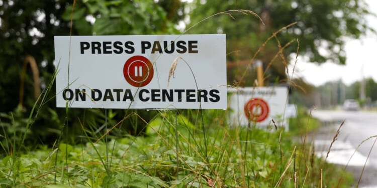 A sign among grass reads PRESS PAUSE ON DATA CENTERS with a red pause button symbol. More similar signs are seen in the background along a roadside.