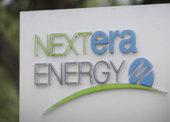 A sign displays the NextEra Energy logo in green, blue, and gray text with a green swoosh and a blue circle featuring a stylized N on a white background. Trees are blurred in the background.