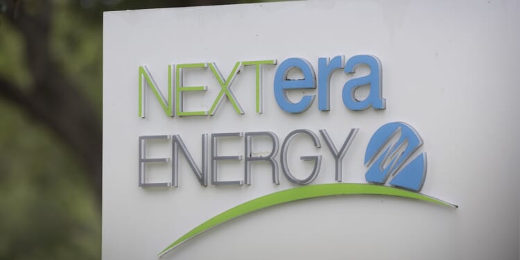 A sign displays the NextEra Energy logo in green, blue, and gray text with a green swoosh and a blue circle featuring a stylized N on a white background. Trees are blurred in the background.