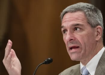 Ken Cuccinelli speaks into a microphone, gesturing with his left hand, appearing to address an audience or panel in a formal setting.