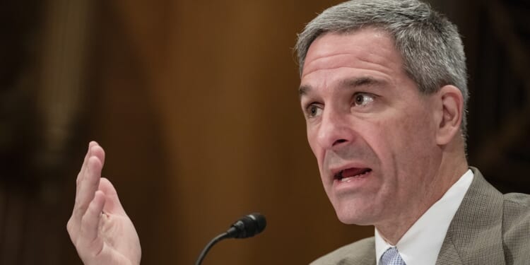 Ken Cuccinelli speaks into a microphone, gesturing with his left hand, appearing to address an audience or panel in a formal setting.