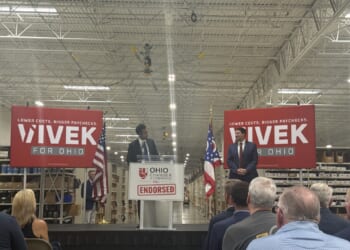 Republican nominee for Ohio governor, Vivek Ramaswamy, looks on at his running mate, Senate President Rob McColley as they stand on a stage in a warehouse, with Vivek for Ohio campaign signs and American and Ohio flags behind them.