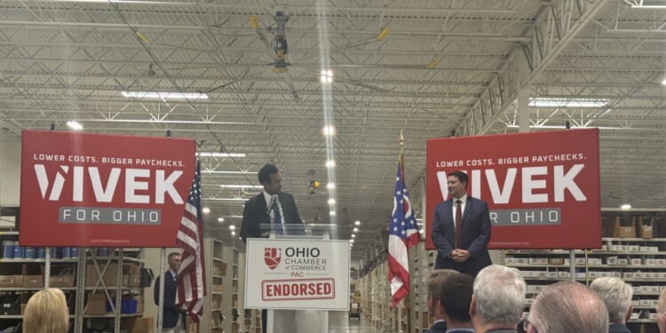 Republican nominee for Ohio governor, Vivek Ramaswamy, looks on at his running mate, Senate President Rob McColley as they stand on a stage in a warehouse, with Vivek for Ohio campaign signs and American and Ohio flags behind them.