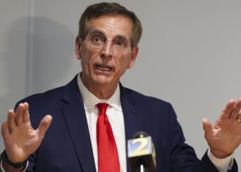A man in a navy suit and red tie speaks with his hands raised in front of microphones at a press conference.