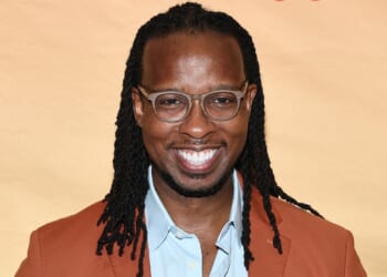 A smiling man with glasses and long braided hair wearing a brown jacket and light blue shirt stands in front of a light tan background.