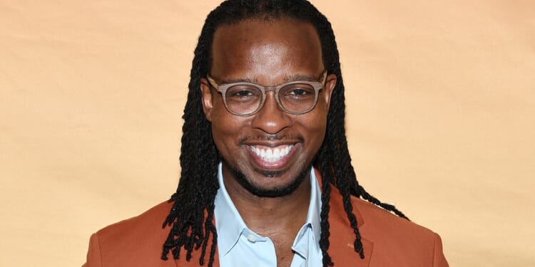 A smiling man with glasses and long braided hair wearing a brown jacket and light blue shirt stands in front of a light tan background.