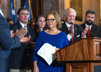 Darline Graham Nordone stands at a podium, surrounded by people applauding her in a formal indoor setting with flags in the background.
