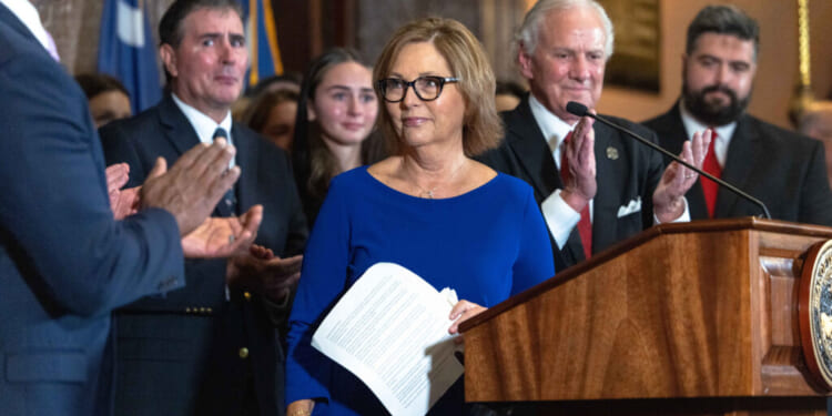 Darline Graham Nordone stands at a podium, surrounded by people applauding her in a formal indoor setting with flags in the background.