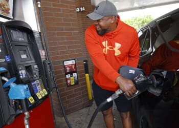 A man in an orange hoodie smiles while pumping gas into a black car at a gas station. He stands next to pump number 4, holding the fuel nozzle.