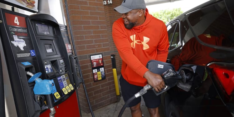A man in an orange hoodie smiles while pumping gas into a black car at a gas station. He stands next to pump number 4, holding the fuel nozzle.