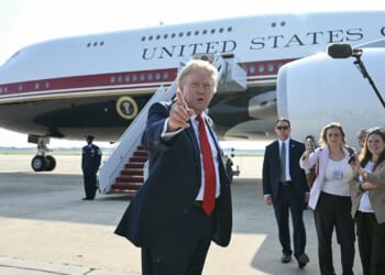 A man in a suit gestures while standing in front of a large plane labeled United States of America, surrounded by security personnel and reporters on an airport tarmac.