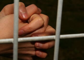 Hands are clasped behind jail bars in a dark cell.