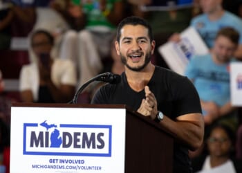 A man speaks enthusiastically at a podium labeled MIDEMS with people in the background holding signs at a public event or rally.