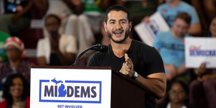 A man speaks enthusiastically at a podium labeled MIDEMS with people in the background holding signs at a public event or rally.