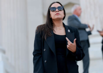 Alexandria Ocasio-Cortez in a black outfit and sunglasses stands confidently outside a building in 2028, gesturing with her hand. Other people are blurred in the background.