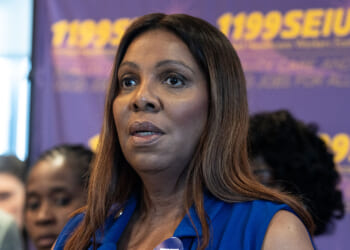 The Justice Department sued Connecticut, New York, and Vermont over their policies giving in-state tuition to illegal aliens. Pictured: New York Attorney General Letitia James, a woman with long brown hair speaks at an event, wearing a blue top. People and a purple and yellow 1199SEIU sign are visible in the blurred background.