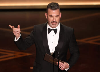 A man in a black tuxedo stands on stage, holding an envelope and gesturing with one hand while speaking into a microphone at a formal event.
