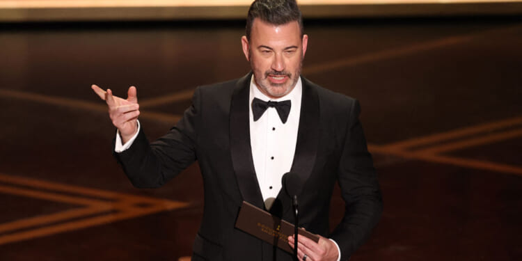 A man in a black tuxedo stands on stage, holding an envelope and gesturing with one hand while speaking into a microphone at a formal event.