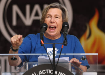 American Federation of Teachers, AFT, President Randi Weingarten. Pictured: a person in a blue shirt speaks passionately at a podium with two microphones, with a sign reading National Action Network and a fiery graphic in the background.