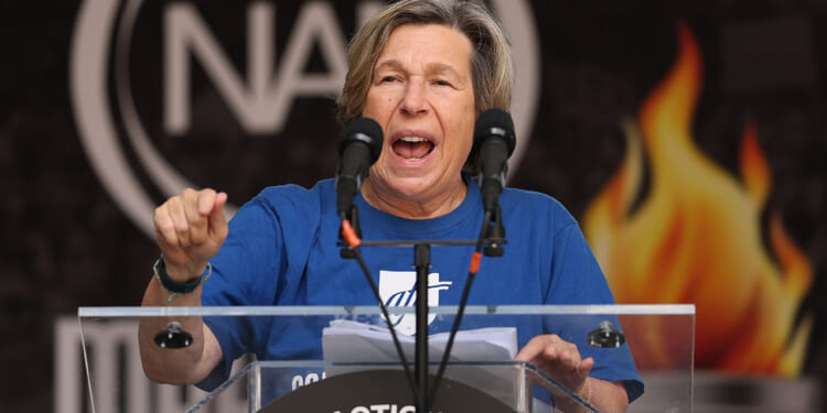American Federation of Teachers, AFT, President Randi Weingarten. Pictured: a person in a blue shirt speaks passionately at a podium with two microphones, with a sign reading National Action Network and a fiery graphic in the background.