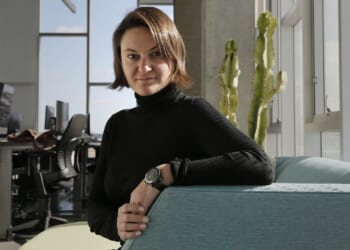 A person with short brown hair, wearing a black turtleneck and watch, sits on a light blue couch in a modern office with computers and a cactus in the background.