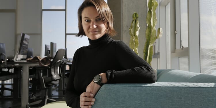 A person with short brown hair, wearing a black turtleneck and watch, sits on a light blue couch in a modern office with computers and a cactus in the background.