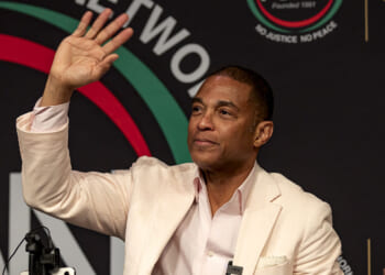 Media outlets have come to the defense of former CNN host Don Lemon in the Cities Church case. Pictured: Lemon, a man in a light-colored suit, sits and waves with his right hand at an event, with a black and colorful backdrop behind him.