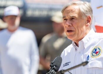 A man in a white shirt with a Texas emblem speaks at a podium outdoors, with people standing in the blurred background.