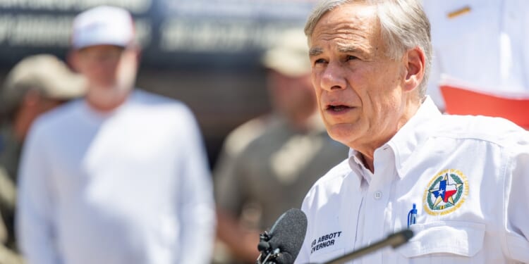 A man in a white shirt with a Texas emblem speaks at a podium outdoors, with people standing in the blurred background.