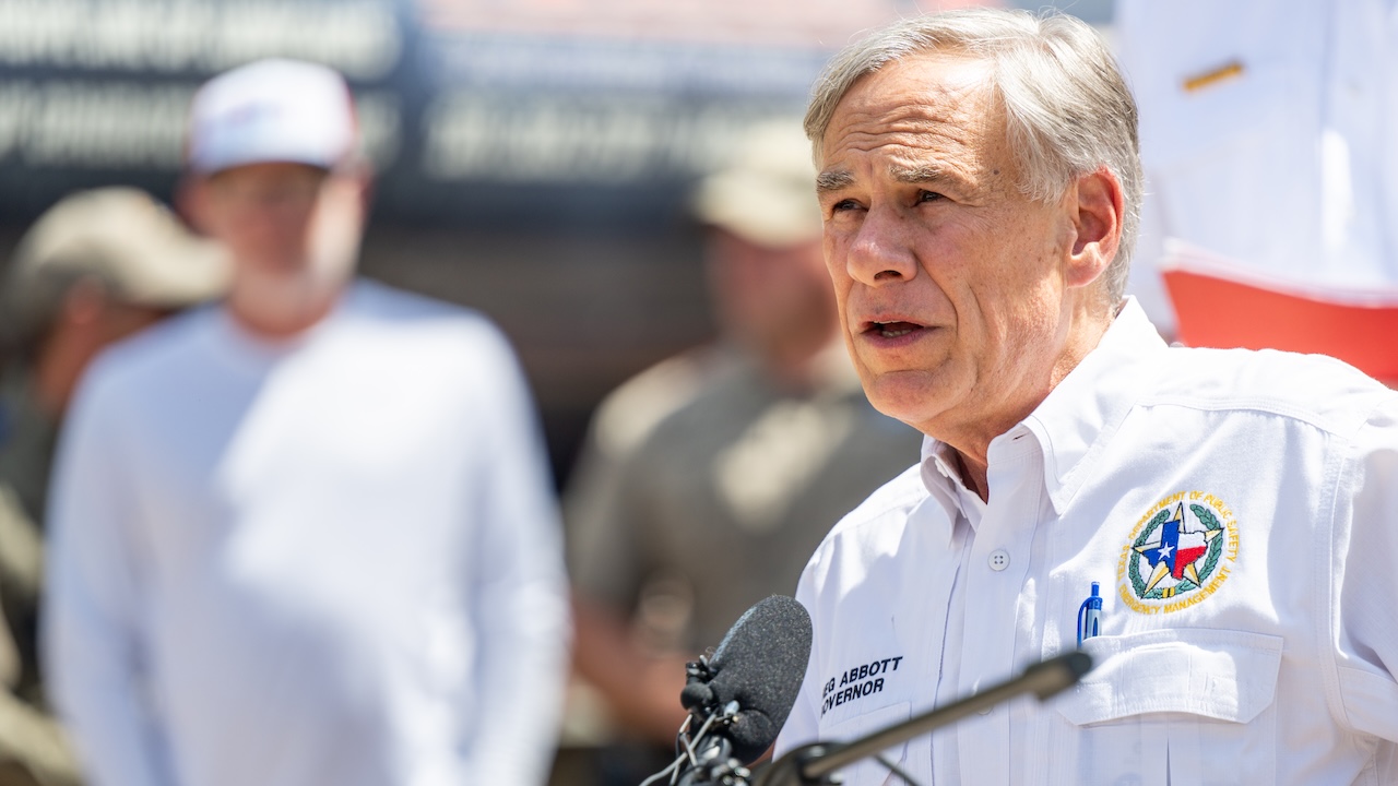 A man in a white shirt with a Texas emblem speaks at a podium outdoors, with people standing in the blurred background.