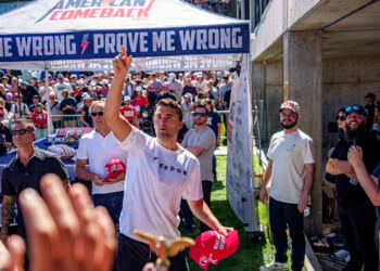 Charlie Kirk stands among a crowd tossing red hats, with people reaching for them. Behind him is a tent with PROVE ME WRONG printed on it. Many in the crowd wear red hats and sunglasses, and the scene is outdoors on a sunny day.