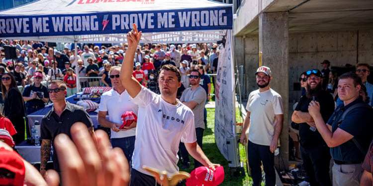 Charlie Kirk stands among a crowd tossing red hats, with people reaching for them. Behind him is a tent with PROVE ME WRONG printed on it. Many in the crowd wear red hats and sunglasses, and the scene is outdoors on a sunny day.