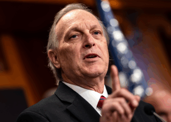 An older man in a suit gestures while speaking at a podium, with an American flag blurred in the background.