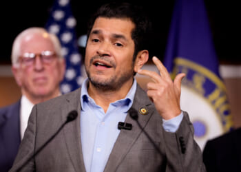 Rep. Jimmy Gomez, in a suit with no tie, speaks at a podium with two other men behind him. U.S. flags and an official emblem are visible in the background.