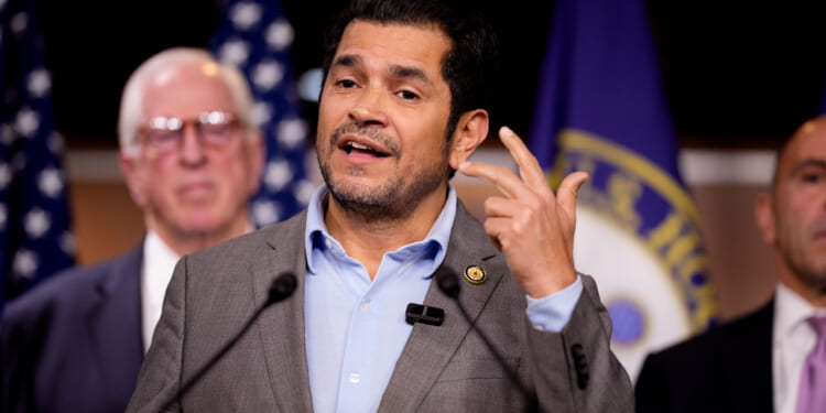 Rep. Jimmy Gomez, in a suit with no tie, speaks at a podium with two other men behind him. U.S. flags and an official emblem are visible in the background.