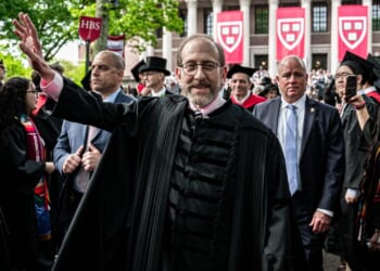 A man in academic robes waves at a graduation ceremony, surrounded by graduates and officials. Harvard banners and a crowd are visible in the background.